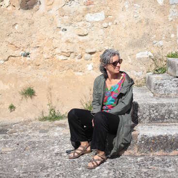 Uma mulher usando óculos escuros, sandálias, uma camisa colorida e uma jaqueta verde está sentada em degraus de pedra contra uma parede áspera, desfrutando pacificamente do ar livre ensolarado.