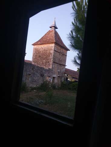 Durch einen dunklen Fensterrahmen sieht man einen rustikalen Steinturm mit spitzem Dach inmitten von Grün, der in sanftes Sonnenlicht getaucht ist und eine malerische, friedliche Atmosphäre ausstrahlt.