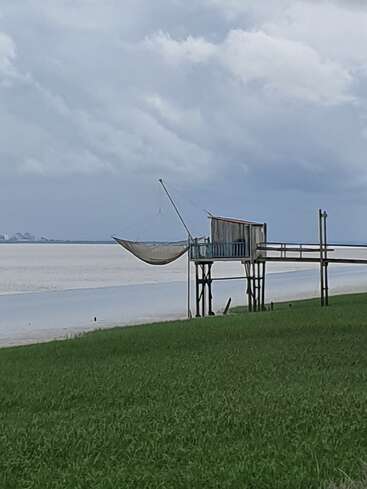 Eine hölzerne Fischerhütte auf Stelzen steht an einem ruhigen, weitläufigen Gewässer mit einem über den Rand hängenden Fischernetz, umgeben von grünem Gras, unter bewölktem Himmel.