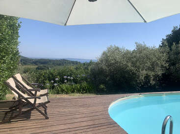 Une scène extérieure relaxante avec deux chaises longues sur une terrasse en bois, un parasol ombragé, une piscine claire, des arbres verdoyants et des collines lointaines sous un ciel bleu.