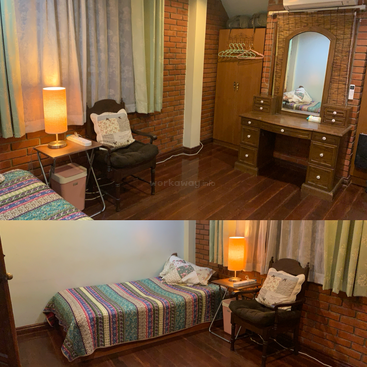 This cozy bedroom features a single bed with a colorful quilt, cushioned chair, side table with lamp, wooden vanity, closet, brick walls, and wooden floors.