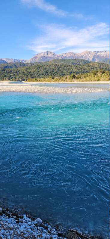 Ein kristallklarer, blauer Fluss fließt sanft an einem steinigen Ufer, einem dichten, grünen Wald und majestätischen Bergen unter einem strahlend blauen Himmel mit Wolkenfetzen darüber vorbei.