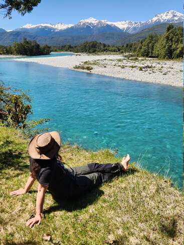 A person in a hat relaxes on grassy riverbank, gazing at turquoise water, lush forest, and majestic snow-capped mountains under a clear blue sky.