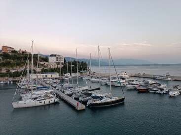 Un puerto deportivo tranquilo al atardecer, repleto de veleros y yates amarrados junto al muelle. Las aguas tranquilas del mar, el cielo sereno y los edificios de la costa completan esta escena de paz.