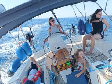 Cuatro personas se relajan en un velero bajo un cielo soleado. Una de ellas lleva el timón, mientras que las demás disfrutan de la brisa y de las vistas del mar, rodeadas de aguas azules y del equipo náutico.