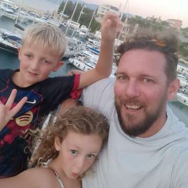 Um homem e duas crianças sorriem para uma selfie em uma marina. Barcos e iates estão atracados ao fundo. Todos parecem felizes e relaxados.