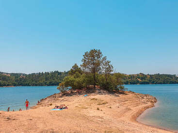 A small sandy peninsula with trees extends into a calm blue lake. People sunbathe, relax, and swim under a clear sky, surrounded by distant greenery.