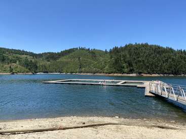 A serene lake scene with a floating dock, people relaxing, surrounded by green hills, clear blue sky, and calm water. Peaceful and inviting natural setting.