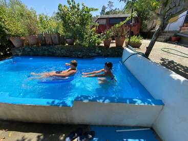 Two children are playing in a small backyard pool on a sunny day. Flower pots, greenery, and a red van are visible in the background.