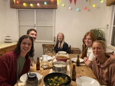 Five people sit around a wooden table, enjoying a cozy meal together. Colorful lights and flags decorate the room, creating a warm, festive atmosphere. Food and drinks abound.