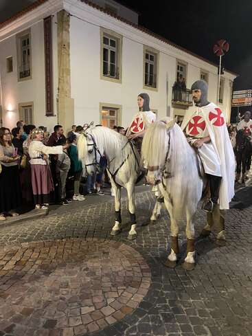 Two men dressed as medieval knights ride white horses in a parade at night, while spectators watch and photograph them on a cobblestone street.