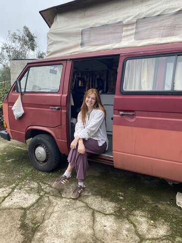 A young woman with red hair smiles, sitting on the step of a red camper van, parked outdoors. The scene feels relaxed and adventurous, evoking travel vibes.