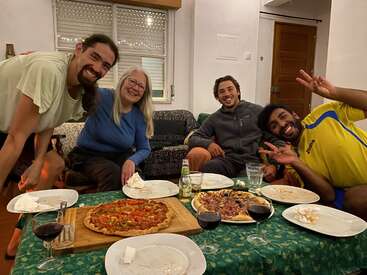 Four friends gather around a table, smiling happily. Homemade pizzas, wine, and drinks are served. The cozy atmosphere suggests a joyful, casual dinner night together.