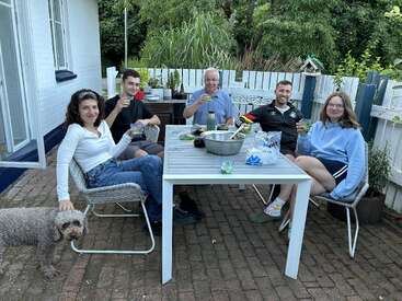 Fünf Leute sitzen um einen Terrassentisch im Freien, lächeln und heben Getränke. Ein Hund mit lockigen Haaren steht in der Nähe. Auf dem Tisch stehen Snacks und Getränke, umgeben von Grünzeug.