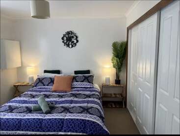 Um quarto aconchegante tem uma cama de casal com roupa de cama estampada, duas mesinhas de cabeceira com abajures, um relógio de parede decorativo, um vaso de planta e portas de armário deslizantes.