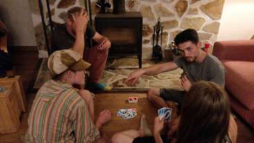 Four friends are sitting on the floor around a wooden table, intensely playing UNO near a cozy fireplace. One player looks frustrated, others are engaged.