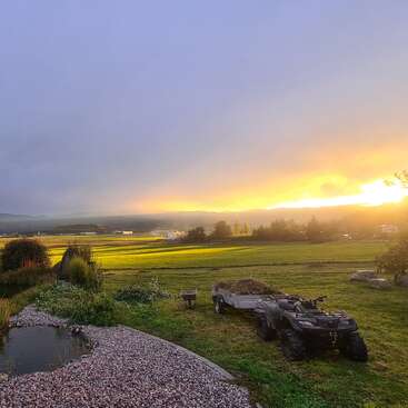 A peaceful countryside scene at sunset, featuring green fields, a pond, a wheelbarrow, a quad bike with a trailer, and distant trees under a colorful sky.