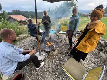 Five friends gather around a smoky outdoor fire pit, grilling food, laughing, and enjoying drinks, surrounded by nature, cozy chairs, and a scenic countryside backdrop.