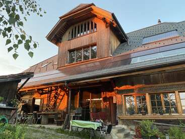 A rustic wooden house with large windows, outdoor seating, and plants. Warm sunlight reflects on the façade, creating a cozy, inviting, and peaceful countryside atmosphere.