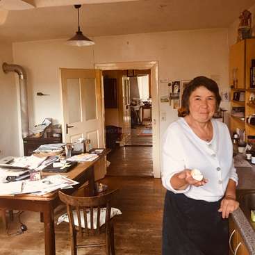 Das Bild zeigt eine Frau, die in einer Küche steht und ein Ei in der Hand hält, mit einem unordentlichen Schreibtisch und einem Holzboden im Hintergrund, umgeben von verschiedenen Gegenständen und Möbeln.