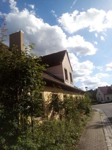 Ein sonniger Tag mit flauschigen Wolken, Pflanzen, die an einem gelben Haus mit Schrägdach wachsen, neben einer ruhigen Straße in einer friedlichen Nachbarschaft.