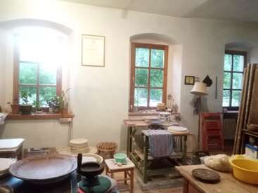 Dieses Bild zeigt eine gemütliche Töpferwerkstatt mit Holzfenstern, Töpferscheiben, gestapelten Keramiken, verschiedenen Werkzeugen, Zimmerpflanzen, natürlichem Licht und einer allgemein kreativen, einladenden Atmosphäre.
