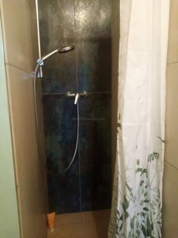 Dieses Bild zeigt eine einfache Dusche mit einem Handbrausekopf, beigen und dunklen Fliesen, einem weißen Duschvorhang mit grünen Mustern und einer orangefarbenen Flasche auf dem Boden.