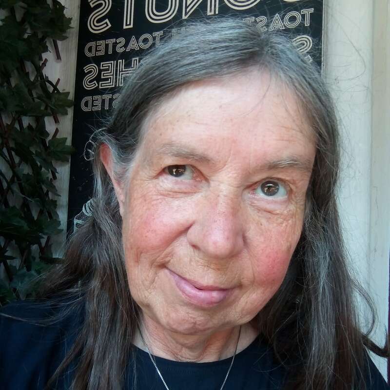 Eine ältere Frau mit langen grauen Haaren lächelt sanft in die Kamera. Sie trägt ein schwarzes Hemd und eine Halskette. Hinter ihr erscheinen Grünpflanzen und ein Schild.