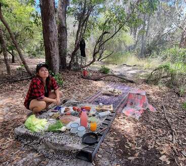 Una mujer sentada en una manta en el bosque, rodeada de un picnic con comida y bebida, con árboles y hojas esparcidas a su alrededor.