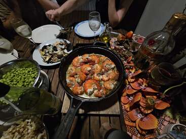 La imagen muestra una mesa de madera cargada con un surtido de platos, incluida una sartén de hierro fundido llena de gambas, en medio de un vibrante mantel de flores y una botella de vino.
