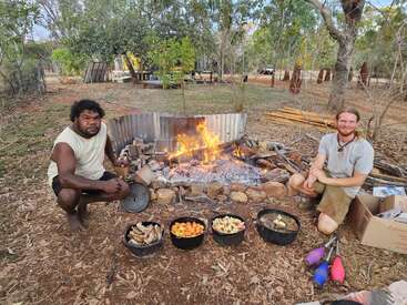 Dos hombres sentados junto a una hoguera, rodeados de ollas con comida, en una zona boscosa con árboles y hierba, disfrutando de una comida juntos.