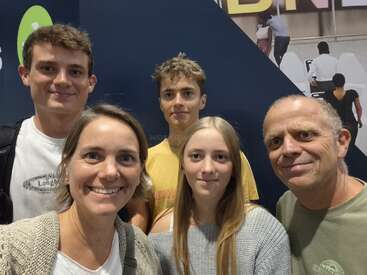 A group of five people, three males and two females, are smiling for a selfie indoors. The background features wall graphics and a partial advertisement.