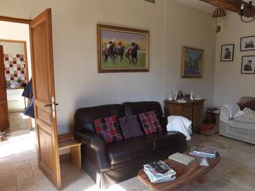 The image depicts a living room with a brown leather couch, a coffee table, and a doorway leading to a kitchen. The walls are white, and the floor is tiled.