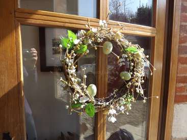 The image depicts a wreath on a wooden door, featuring white flowers and green leaves, with a person\'s reflection visible in the glass.