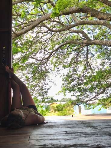 Une personne est allongée sur un sol en bois, les jambes relevées contre une porte, sous de grandes branches d'arbre, profitant de la lumière du soleil et d'une vue paisible sur la nature.