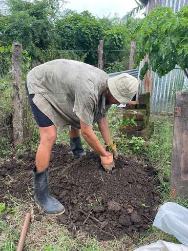 Une personne portant un chapeau, des gants et des bottes creuse ou plante dans un jardin. Elle est entourée de verdure, de poteaux en bois et d'un mur en tôle ondulée.