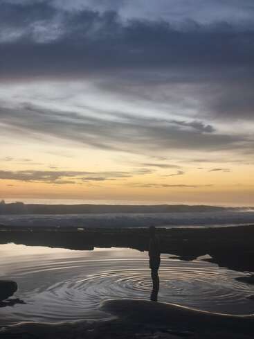 Une personne se tient dans une eau peu profonde au coucher du soleil, entourée de douces ondulations. Le ciel est spectaculaire, parsemé de nuages, et l'horizon s'illumine doucement. Ambiance paisible.