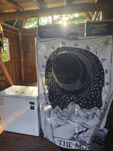 Cette image montre une pièce rustique en bois avec un congélateur, deux grandes batteries et une tapisserie mystique représentant une lune, des étoiles, des montagnes et des symboles du zodiaque.