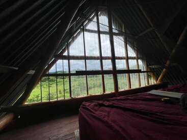 Une chambre mansardée confortable avec des poutres en bois, une fenêtre triangulaire et un lit recouvert d'un drap rouge donne sur un paysage verdoyant et un ciel nuageux à l'extérieur.