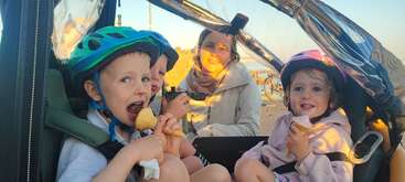 Das Bild zeigt eine Gruppe von Kindern, die in einer Reihe sitzen und alle die gleiche Kleidung tragen, während eine große Gruppe von Erwachsenen hinter ihnen steht. Die Kinder halten Bücher in der Hand und scheinen sich in einer Schulumgebung zu befinden.