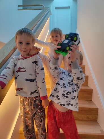 Das Bild zeigt drei Kinder, die auf einer Treppe stehen. Zwei halten Spielzeug in der Hand und eines lehnt am Geländer, alle tragen Pyjamas.