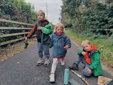 Drei Kinder gehen einen gepflasterten Weg entlang, wobei ein Kind auf dem Boden sitzt, umgeben von einem Holzzaun und Bäumen, in einer Außenanlage.