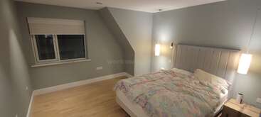 Das Bild zeigt ein Zimmer mit einem Bett, einer Kommode und einem Nachttisch, mit einer weißen Bettdecke und Kissen, vor einer beigen Wand mit einem Fenster und einer Tür. Der Raum ist gut beleuchtet und aufgeräumt.