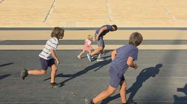 Four children and one adult are racing on a track under bright sunlight. The background shows large empty bleachers, emphasizing their energetic movement and playful competition.