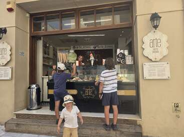 Three children stand outside an open-air gelato shop called Arte Athens, choosing ice cream flavors while a woman behind the counter smiles at them.