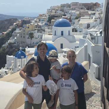 A family of five poses in front of picturesque white buildings with blue domes in Santorini, Greece, under a clear sky. The sea is visible.