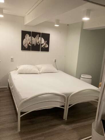 Un dormitorio sencillo con una cama doble con sábanas blancas y dos almohadas, obras de arte de orquídeas encima de la cama, suelo de madera clara y decoración moderna minimalista.