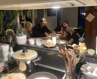 Cuatro personas se sientan alrededor de una mesa de cocina, conversando y dibujando. La encimera de la cocina, en primer plano, contiene utensilios, cactus, velas y alimentos para cocinar.