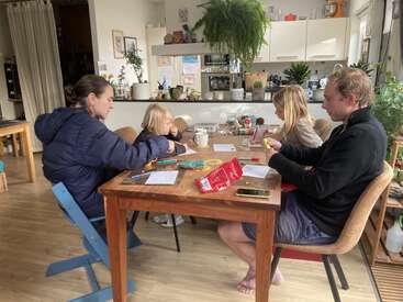 Cuatro personas se sientan alrededor de una mesa de madera en una acogedora cocina llena de plantas, dedicadas a actividades artísticas con rotuladores, papel y bocadillos. El ambiente parece relajado y creativo.