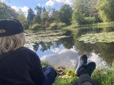 Dos personas se relajan junto a un sereno estanque, rodeadas de frondosos árboles verdes y nenúfares, bajo un cielo azul lleno de esponjosas nubes blancas, disfrutando de la naturaleza.